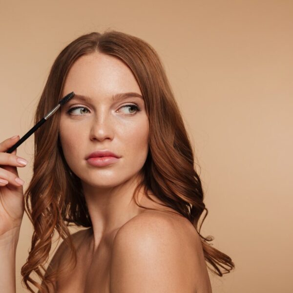Beauty portrait of calm ginger woman with long hair looking away and posing sideways while combing her eyebrows with brush over cream background